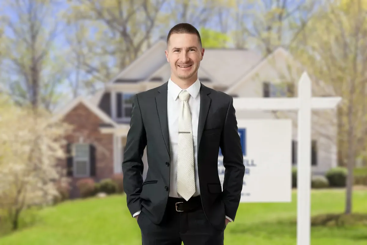 Justin Paupst-Wall in a dark suit, hands in his pockets, in front of a house with a For Sale sign